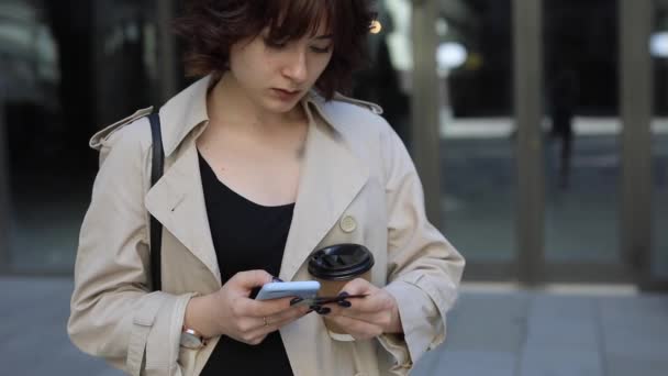 Мила жінка спілкується в смартфоні в траншеї пальто — стокове відео