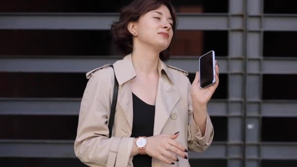 Мила жінка спілкується в смартфоні в траншеї пальто — стокове відео