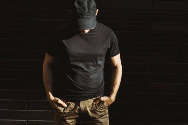 Siyah tişörtlü ve beyzbol şapkalı adam siyah duvara karşı poz veriyor.