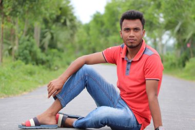 Kot pantolon, tişört ve slayt terlik giyen şık genç Asyalı bir adam bir caddenin ortasında oturup poz veriyor.