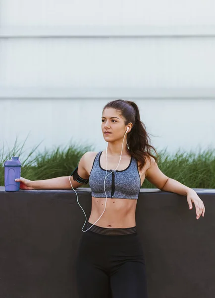 Gri arka planı ve parlak duvara yeşil çim üzerinde egzersiz yaptıktan sonra dinlenme sportif kadın. Uygun egzersiz yaptıktan sonra kız giyiyor sportif ilk ve müzik dinleme. Sağlıklı yaşam ve fitness kavramı