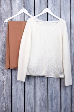 Beyaz kazak ve kahverengi pantolon askıları üzerinde.