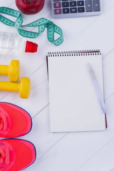 Healthy and active lifestyle concept. Sport shoes, dumbbells, bottle of water, measuring tape, apple, calculator and blank notepad. Sport fitness background.