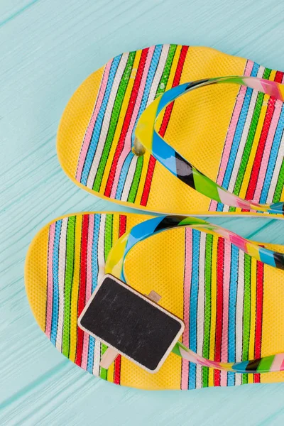 Helle Strand-Flip-Flops mit kleiner leerer Tafel auf azurblauem Hintergrund. — Stockfoto