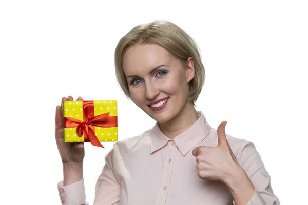 Feliz mujer rubia sonriente sosteniendo caja de regalo y haciendo buen gesto . — Foto de Stock