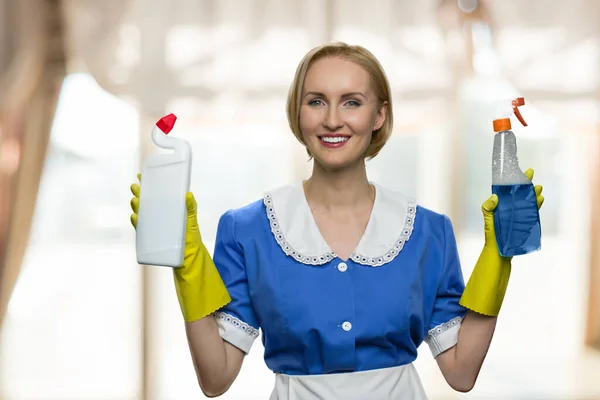 Linda empregada doméstica segurando suprimentos de limpeza e olhando para a câmera. — Fotografia de Stock