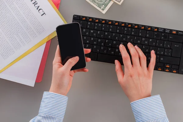 Female hands using smartphone while typing on computer keyboard.