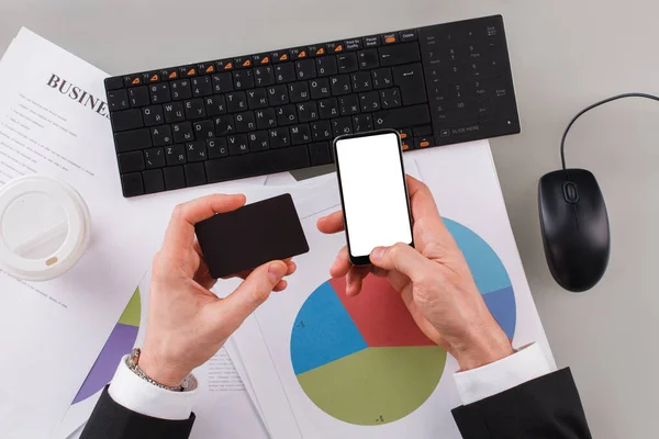 Male hands holding business card and mobile phone in office.