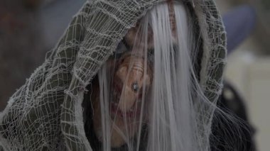 Uzun burunlu, beyaz saçlı ve yırtık pırtık bir gazlı bez pelerinli kapüşonlu cadılar bayramı cadısının yakın çekimi. Banliyö evindeki ürkütücü bahçe dekorasyonu ürkütücü bir ruh hali yaratıyor..