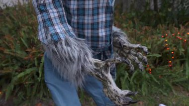 Gri kürklü ve keskin dişli hırıldayan kurt adam maskesi. Kostüm karakteri pençeli elleri açık havada, mavi ekose gömlekli bir bahçede gösteriyor..