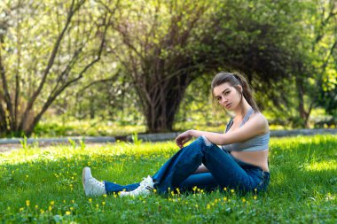 Güzel bir genç kız güneşli bir günde yeşil çimlerin üzerinde oturur. Açık havada. Doğanın tadını çıkar. Baharda çimlerin üzerinde gülümseyen sağlıklı bir kız. Bahar yaşam tarzı