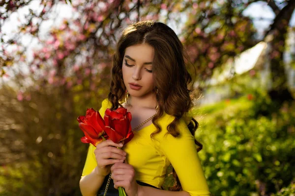 若いです魅力的な女の子でA黄色のドレス保持A花束の赤いチューリップでザ庭 — ストック写真