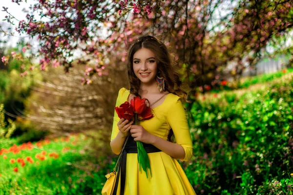 波状の茶色の髪をした美しい若い女の子は 赤い花を咲かせる植物園に立っています 自然なメイク 黄色のドレス ファッション — ストック写真