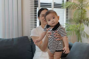 Genç bir anne, bebeğini sevgiyle kucaklar ve bir kanepede emzirir. Erken çocukluk ve aile hayatının anne bakımını, bağlarını ve besleyici anlarını sembolize eder..