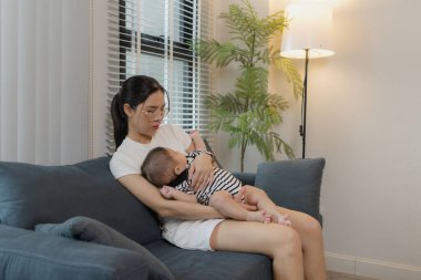 Genç bir anne, bebeğini sevgiyle kucaklar ve bir kanepede emzirir. Erken çocukluk ve aile hayatının anne bakımını, bağlarını ve besleyici anlarını sembolize eder..