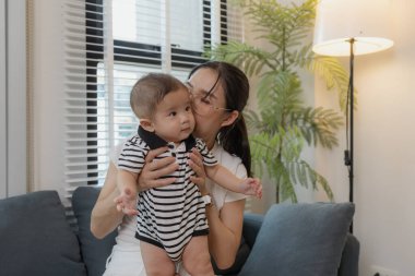 Genç bir anne, bebeğini sevgiyle kucaklar ve bir kanepede emzirir. Erken çocukluk ve aile hayatının anne bakımını, bağlarını ve besleyici anlarını sembolize eder..