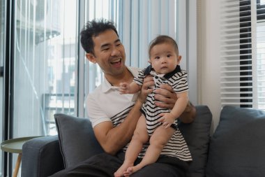 Neşeli bir baba bebeğini kollarında tutuyor, evdeki kanepede oturuyor. Bebek meraklı ve sakin görünürken baba sıcak bir gülümsemeyle aile bağlarını yakalıyor..