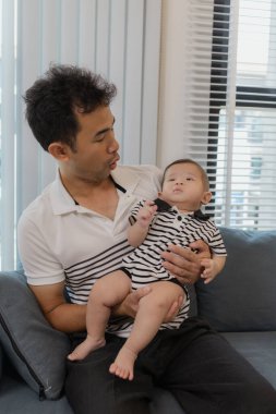 Neşeli bir baba bebeğini kollarında tutuyor, evdeki kanepede oturuyor. Bebek meraklı ve sakin görünürken baba sıcak bir gülümsemeyle aile bağlarını yakalıyor..