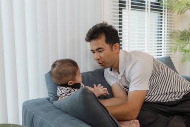 Neşeli bir baba, bebeğiyle oturma odasında bağ kuruyor. Bebek ellerini tutarken sıcak bir şekilde gülümsüyor. Sevgi, neşe ve ebeveynliğin mutluluğunu yansıtan an..