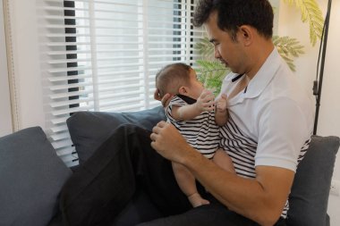 Neşeli bir baba, bebeğiyle oturma odasında bağ kuruyor. Bebek ellerini tutarken sıcak bir şekilde gülümsüyor. Sevgi, neşe ve ebeveynliğin mutluluğunu yansıtan an..