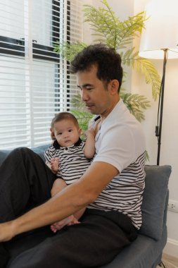 Neşeli bir baba, bebeğiyle oturma odasında bağ kuruyor. Bebek ellerini tutarken sıcak bir şekilde gülümsüyor. Sevgi, neşe ve ebeveynliğin mutluluğunu yansıtan an..