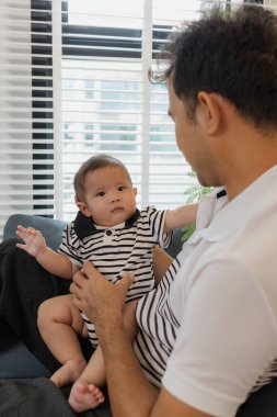 Neşeli bir baba, bebeğiyle oturma odasında bağ kuruyor. Bebek ellerini tutarken sıcak bir şekilde gülümsüyor. Sevgi, neşe ve ebeveynliğin mutluluğunu yansıtan an..