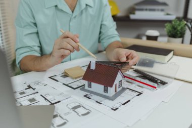 Bir mimar mimari planlara yerleştirilmiş bir ev modelini ölçmek için üçgen bir cetvel ve kalem kullanır, tasarım süreci sırasında oranları ve detayları kontrol eder.