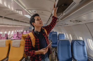 Genç adam eşyalarını bir uçağın üst kompartımanına koyarak bir uçuş için hazırlanıyor. Neşeli ve seyahat macerasına hazır görünüyor, elinde bir sırt çantası ve pasaportuyla..