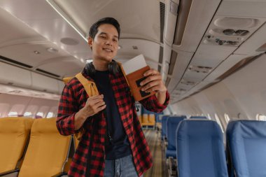 Asyalı bir adam uçağa biniyor ve elinde pasaport ve uçuş kartıyla koltuğunu arıyor. Sırt çantası taşırken ve boynunda kulaklık varken mutlu ve heyecanlı görünüyor..