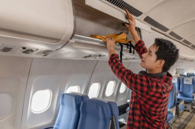 Genç adam eşyalarını bir uçağın üst kompartımanına koyarak bir uçuş için hazırlanıyor. Neşeli ve seyahat macerasına hazır görünüyor, elinde bir sırt çantası ve pasaportuyla..