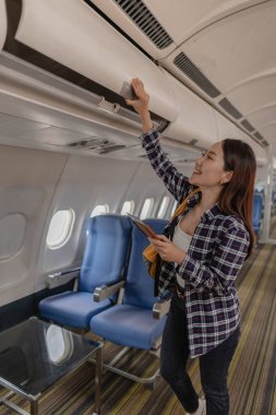 Gülümseyen genç bir kadın, pasaportunu ve uçuş kartını tutarken başınızın üstündeki kompartımanı açmaya çalışıyor. Gelecek yolculuğuna hazırlanırken neşeli ve heyecanlı görünüyor..
