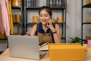 Gülümseyen genç bayan girişimci cep telefonuyla konuşuyor ve ev ofisinden dizüstü bilgisayarla çalışıyor, çevrimiçi giyim mağazasının müşteri siparişlerini yönetiyor ve sevkiyat ve teslimat için ürünler hazırlıyor..