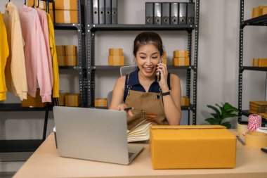 Gülümseyen genç bayan girişimci cep telefonuyla konuşuyor ve ev ofisinden dizüstü bilgisayarla çalışıyor, çevrimiçi giyim mağazasının müşteri siparişlerini yönetiyor ve sevkiyat ve teslimat için ürünler hazırlıyor..