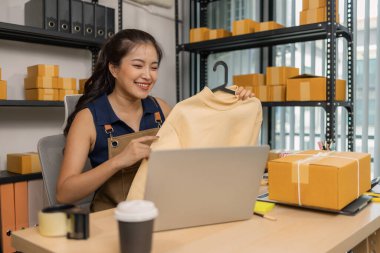 Genç bayan girişimci, ev ofisinden kıyafet satmak için yaşarken gülümsüyor. Moda ürünleri sunar, izleyicilerle iletişim kurar ve canlı seans sırasında online mağazasını yönetir..