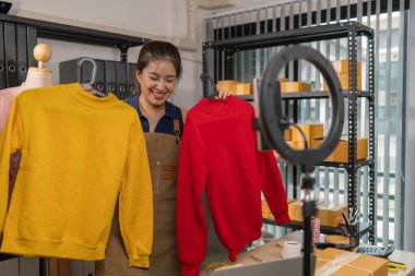 Genç bayan girişimci, ev ofisinden kıyafet satmak için yaşarken gülümsüyor. Moda ürünleri sunar, izleyicilerle iletişim kurar ve canlı seans sırasında online mağazasını yönetir..