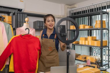 Genç bayan girişimci, ev ofisinden kıyafet satmak için yaşarken gülümsüyor. Moda ürünleri sunar, izleyicilerle iletişim kurar ve canlı seans sırasında online mağazasını yönetir..