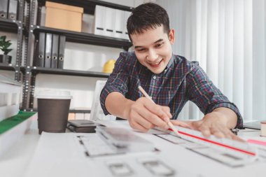 Ofis masasında çalışan genç erkek mimar, inşaat planları çiziyor ve tasarım araçları kullanıyor, mimaride yaratıcılık ve adanmışlık gösterirken tasarım üzerine odaklanıyordu..