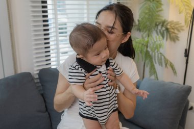 Genç bir anne, bebeğini sevgiyle kucaklar ve bir kanepede emzirir. Erken çocukluk ve aile hayatının anne bakımını, bağlarını ve besleyici anlarını sembolize eder..