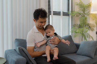 Neşeli bir baba, bebeğiyle oturma odasında bağ kuruyor. Bebek ellerini tutarken sıcak bir şekilde gülümsüyor. Sevgi, neşe ve ebeveynliğin mutluluğunu yansıtan an..