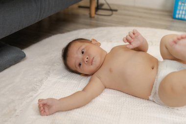 Bebek bezi içinde yumuşak beyaz bir battaniyeye uzanmış kollarını hareket ettirirken rahatça dinleniyor. Bebek sakin ve meraklı görünüyor, masumiyeti ve çocukluk anılarını yakalıyor..