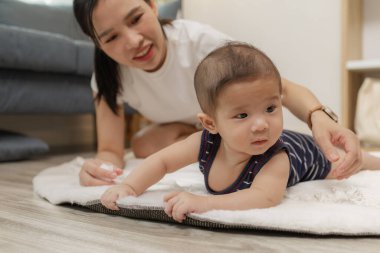 Anne sıcak bir gülümsemeyle onu cesaretlendirirken, bir bebek yumuşak bir paspasın üzerinde midede vakit geçirmekten zevk alır. Bu hassas an, erken gelişimi, aile bakımını ve evde kaynaşmayı vurguluyor.