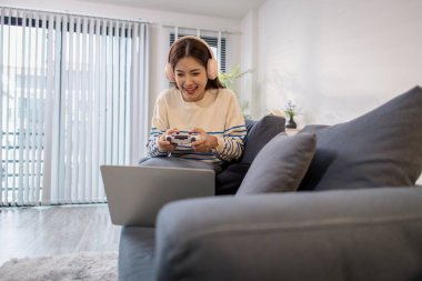 Genç bir kadın, elinde oyun kumandası ve dizüstü bilgisayarıyla video oyunu oynarken kulaklık takarak koltukta oturuyor. Neşeli ifadesi neşe ve heyecan gösteriyor..