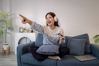 Genç bir kadın, elinde oyun kumandası ve dizüstü bilgisayarıyla video oyunu oynarken kulaklık takarak koltukta oturuyor. Neşeli ifadesi neşe ve heyecan gösteriyor..