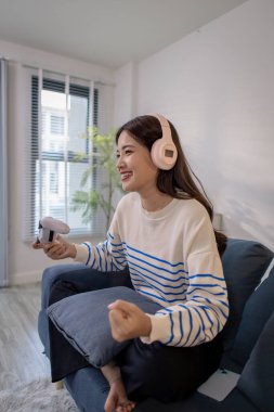 Genç bir kadın, elinde oyun kumandası ve dizüstü bilgisayarıyla video oyunu oynarken kulaklık takarak koltukta oturuyor. Neşeli ifadesi neşe ve heyecan gösteriyor..