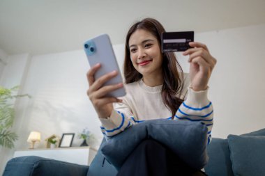 Bir elinde akıllı telefon, diğer elinde kredi kartı, dijital finans ve e-ticaret yaşam tarzını temsil eden bir genç kadın, evinde bir kanepede oturmuş gülümsüyor..