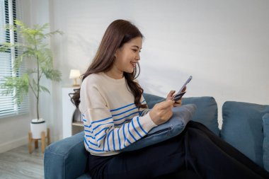 Bir elinde akıllı telefon, diğer elinde kredi kartı, dijital finans ve e-ticaret yaşam tarzını temsil eden bir genç kadın, evinde bir kanepede oturmuş gülümsüyor..