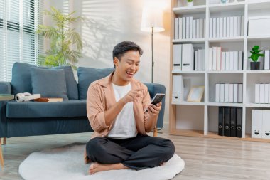 Genç bir adam, rahat bir oturma odasında akıllı telefon kullanırken bacak bacak bacak üstüne atmış, gülümseyerek yerde rahatça oturuyor. Rahatlamış atmosfer konfor, yaşam tarzı ve ev bağlantısını vurguluyor.