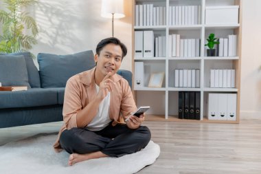 Genç bir adam, rahat bir oturma odasında akıllı telefon kullanırken bacak bacak bacak üstüne atmış, gülümseyerek yerde rahatça oturuyor. Rahatlamış atmosfer konfor, yaşam tarzı ve ev bağlantısını vurguluyor.