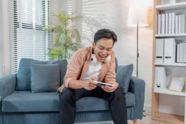 Genç bir adam evinde bir koltuğa oturmuş, kulaklıkları boynunda, akıllı bir telefon tutuyor ve mobil bir oyun oynuyor, gülümsüyor ve heyecan ve mutlulukla kutluyor..