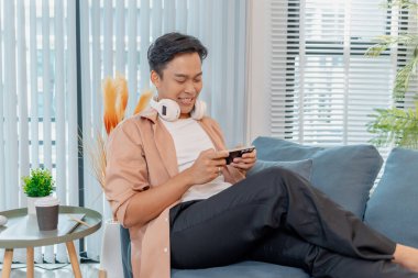 Genç bir adam evdeki kanepede oturuyor, kulaklıkları boynunda, günlük kıyafetlerle, gülümsüyor ve kutluyor, akıllı telefonuna bakıyor, neşe ve heyecan gösteriyor..